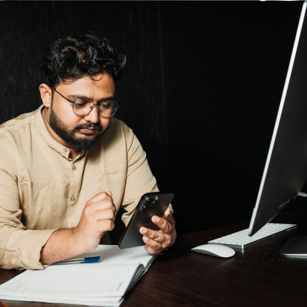 man sitting I a dark room looking at a black phone in his hand while his arm rests on top of.a notebook