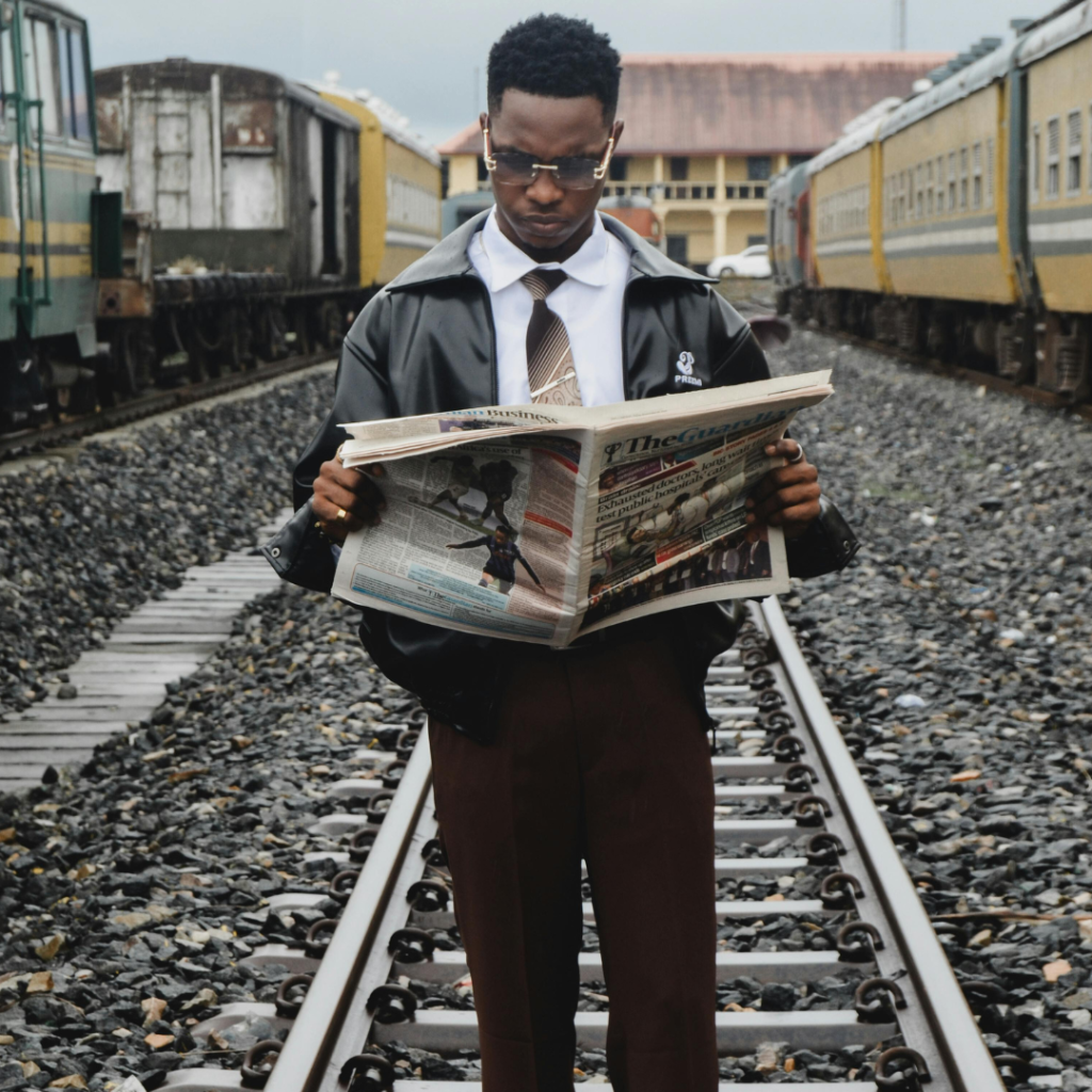 Man wearing a tie and glasses standing on a raildroad track holding a newspaper