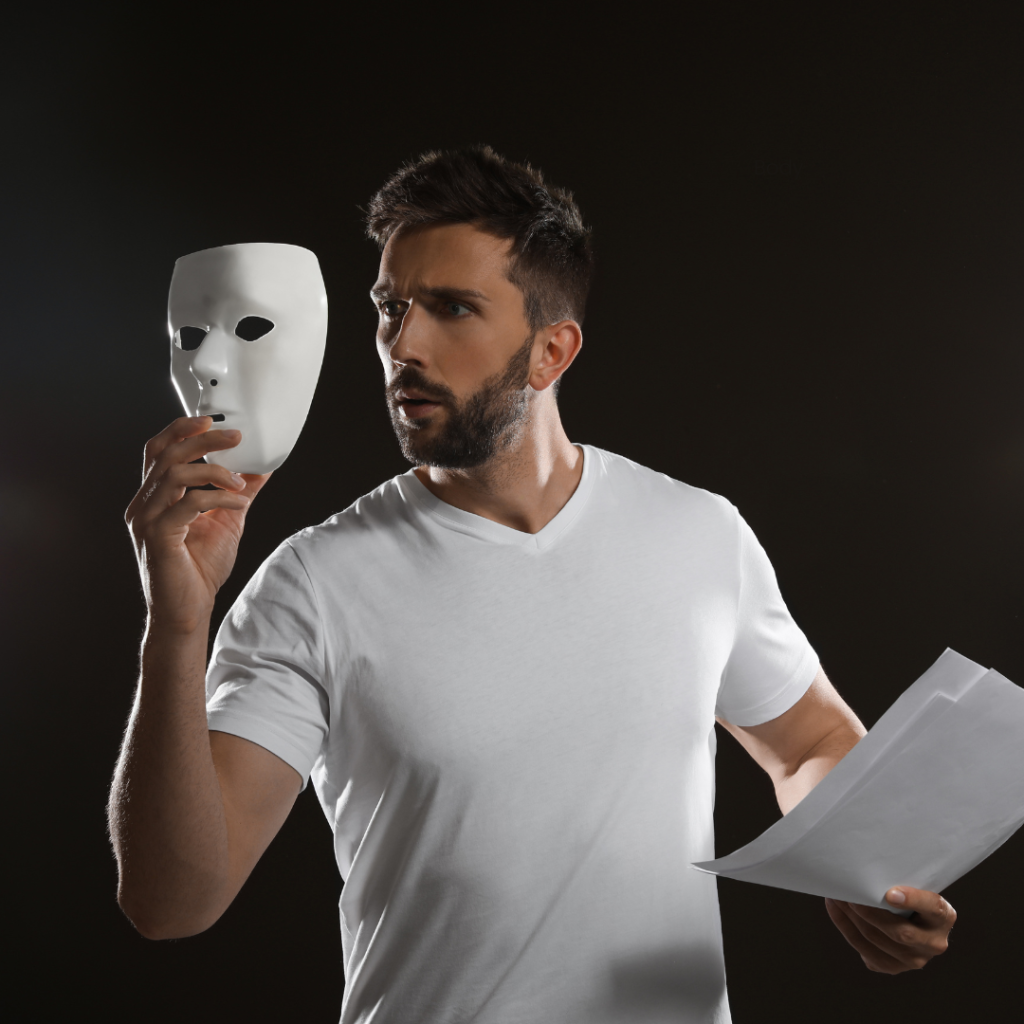 Man in a white T-Shirt standing in front of a black back round holding a theatre mask
